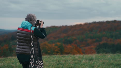 Cinematic-shot-of-a-woman-photographer-with-grey-hair-taking-pictures-of-an-autumn-landscape-with-her-camera-during-a-windy-day-surrounded-by-nature-in-slow-motion