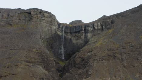 Toma-Aérea-Acercándose-A-Una-Cascada-Natural-En-Una-Montaña-Rocosa-En-Islandia-Durante-El-Día-Nublado