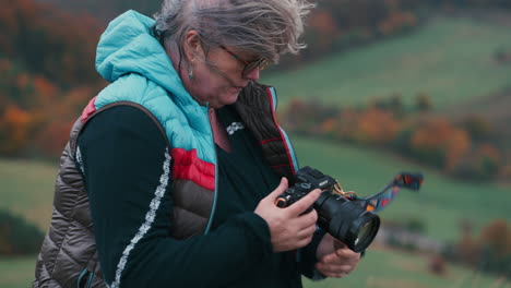 Close-up-shot-of-a-woman-photographer-looking-at-her-camera,-smiling-and-taking-pictures,-wind-playing-with-her-grey-hair-in-an-autumn-landscape-with-colourful-orange-leaves