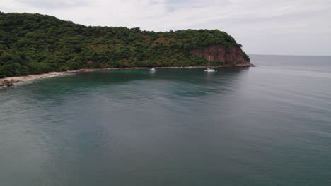 Frontal-drone-flight-from-sea-to-Punta-de-Mita's-rocky-shores,-with-boats-and-a-lush-green-mountain-contrasted-by-blue-sea