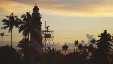 Rack-focus-to-a-beautiful-old-lighthouse-at-sunset-in-Kerala-with-coconut-trees,-birds-flying-past-and-a-radar-tower-spinning-beside