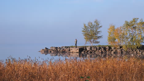 Man-enjoys-autumn-weather-on-lake-coastline-in-Scandinavia,-distance-view
