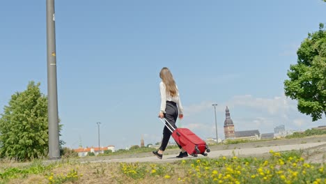Woman-walking-at-the-park-while-pulling-along-a-wheeled-luggage-bag,-handheld-tracking-shot