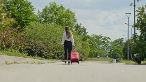 Woman-walking-on-the-sidewalk-while-pulling-along-a-wheeled-luggage-bag,-handheld-wide-shot