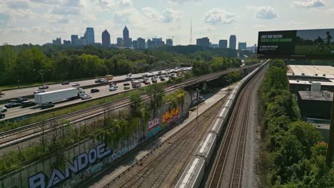 Aerial-view-of-stopped-train-on-railroad-beside-busy-highway-and-skyline-of-Atlanta-City-in-backdrop