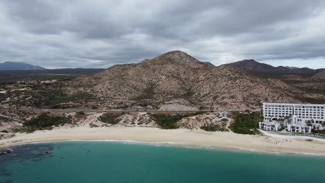 Aerial-backwards-shot-of-the-beautiful-coastal-landscape-in-los-cabos-with-a-view-of-the-sandy-beach,-a-busy-coastal-road-and-the-turquoise-sea-and-the-renowned-Marquis-hotel-during-a-mexico-vacation