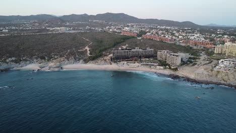 Aerial-view-over-the-coastline-from-monument-beach-at-the-cape-thompson-hotel-in-los-cabos-overlooking-the-boardwalk,-blue-sea-with-calm-waves-and-the-hotel-grounds-during-a-vacation-in-mexico
