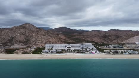 Aerial-panning-shot-in-front-of-the-luxurious-hotel-of-the-Marquis-Los-Cabos-Hotel-with-a-view-of-the-coastal-landscape-with-a-dreamlike-beach,-turquoise-sea-water-and-majestic-mountains-in-background
