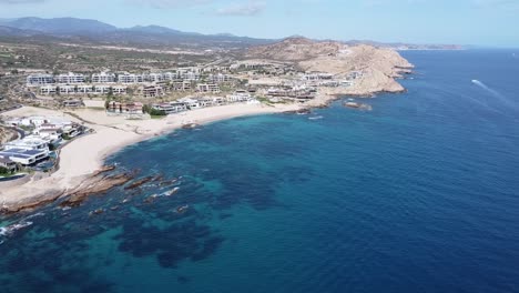 Aerial-view-over-the-picturesque-coast-of-Playa-Boca-del-Tule-in-baja-california-sur-with-a-view-of-the-blue-sea,-various-hotel-complexes-and-majestic-mountains-in-the-background
