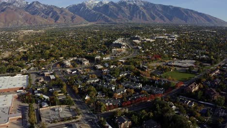 Tilt-up-descending-aerial-drone-wide-landscape-shot-revealing-the-stunning-snowcapped-rocky-mountains-of-Utah-with-Salt-Lake-county-below-full-of-buildings-and-colorful-trees-on-a-warm-sunny-fall-day