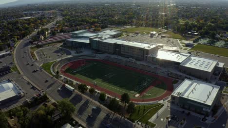Dolly-out-tilt-up-aerial-drone-shot-of-a-large-modern-American-high-school-and-the-large-empty-track-and-football-field-next-to-a-busy-street-on-a-warm-sunny-fall-day-in-Salt-Lake-County,-Utah