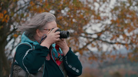Female-Photographer-with-grey-hair-and-glasses-taking-pictures-with-her-camera-in-nature,-surrounded-by-orange-leaved-trees-during-a-cold-windy-autumn-day-in-slow-motion