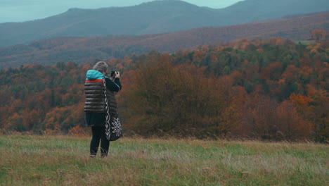 Older-female-Photographer-with-grey-hair-taking-pictures-with-her-camera-in-nature-surrounded-by-autumn-coloured-trees-and-grass-during-a-cold-windy-day-in-slow-motion