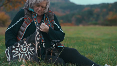Fat-older-woman-with-grey-hair-and-camera-looking-for-another-lens-in-her-bag-while-sitting-on-the-grass-in-nature-during-a-windy-autumn-day-surrounded-by-colourful-trees-in-slow-motion