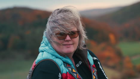 Close-up-shot-of-a-smiling-fat-woman-with-grey-hair-wearing-glasses-and-colourful-warm-vest,-surrounded-by-beautiful-autumn-nature-with-orange-trees-in-the-background-during-a-cold-windy-sunset