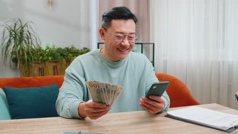 Rich-happy-Asian-man-counting-money-cash-on-smartphone-calculator-app,-calculate-income-earnings