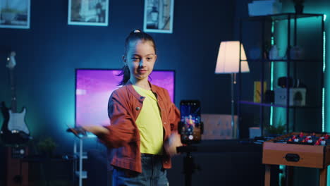 Kid-dancing-in-dimly-lit-home-studio-interior,-producing-content-with-smartphone-on-tripod