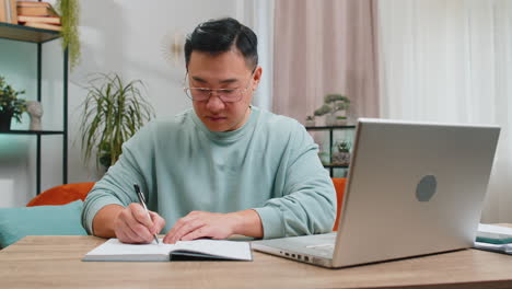 Hombre-Asiático-Independiente-Tomando-Notas-En-Un-Cuaderno-En-Casa,-Usando-Una-Computadora-Portátil,-Preparando-Un-Informe-Financiero-Empresarial