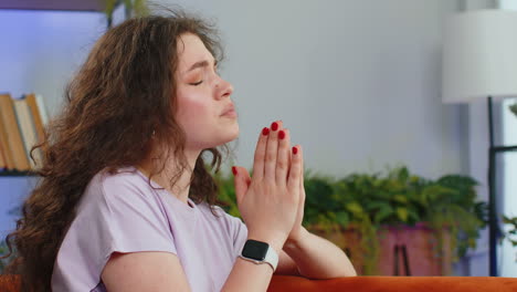 Young-woman-girl-praying-sincerely-with-folded-arms-asking-God-for-help-begging-apology-on-home-sofa