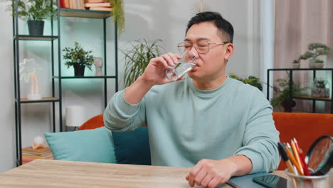 Thirsty-Asian-man-sits-at-home-table-holding-glass-of-natural-aqua-make-sips-drinking-filtered-water