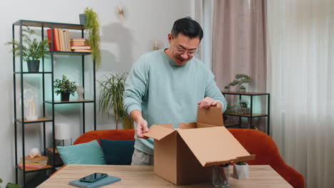 Happy-Chinese-man-shopper-unpacking-cardboard-box-delivery-parcel-online-shopping-purchase-at-home