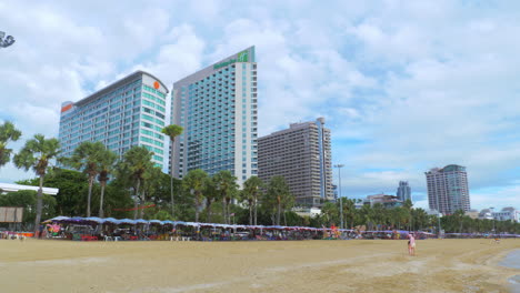 Strand-Vor-Drei-Hochhaushotels-Mit-Weißem-Sand-Und-Einer-Reihe-Von-Stranddächern-Oder-Sonnenschirmen,-Die-Von-Menschen-Und-Kindern-Besetzt-Sind
