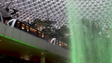 Skytrain-Going-Past-Overhead-Beside-Falling-Rain-Vortex-Waterfall-With-Green-Light-Glow-At-Jewel-Changi-Airport