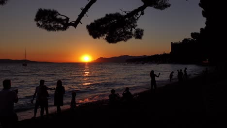 Dunkle-Silhouetten-Von-Menschen-Und-Familien,-Die-Am-Strand-In-Makarska-In-Kroatien-Spielen,-Mit-Wunderschönem-Sonnenuntergang-Im-Hintergrund