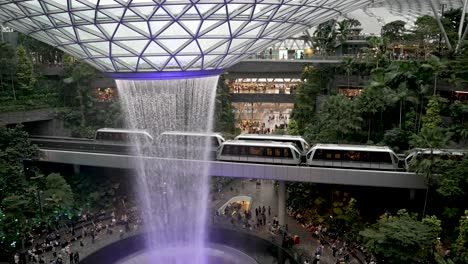 High-Angle-View-Overlooking-Purple-Illuminated-Indoor-Waterfall-At-Jewel-Changi-Airport-Singapore-With-Skytrains-Passing-Each-Other
