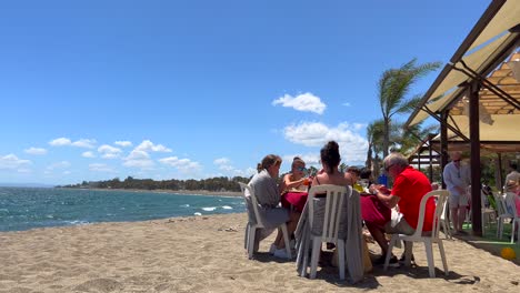 Family-sitting-and-eating-at-a-beach-restaurant-on-a-very-windy-day-in-San-Pedro-de-Alcantara,-sunny-summer-day-in-Marbella-Spain,-4K-shot