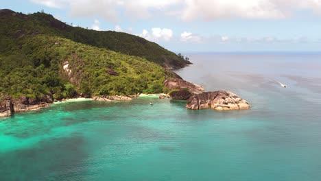 Drone-shot-of-passing-boat,-lush-mountain-with-huge-granite-stone,-Mahe,-Seychelles-60-fps