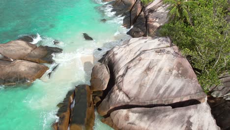Reveal-drone-shot-of-white-sandy-beach,-large-granite-stone,-trees-and-waves-crashing-on-the-shore,-Mahe-Seychelles-60-fps