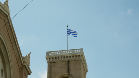 Greek-flag-waving-on-historic-building's-tower-against-blue-sky