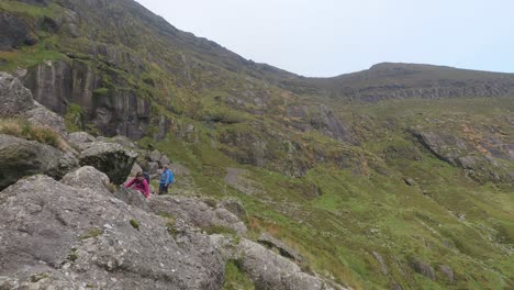 Girls-climbing-over-rocks-with-panning-shot-to-Coumshingaun-Lake-high-in-the-Comeragh-Mountains-Waterford-Ireland-on-a-perfect-winter-day