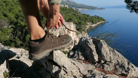 Hiker-man-on-top-of-mountain-cliff-edge-with-sea-view-tying-shoe-laces,-close-up