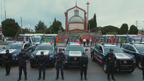 Vertical-pan-of-7-police-pick-ups-and-2-ambulances-with-armed-officers-of-the-Police-Department-lined-up-and-posing-in-front-of-their-cars,-with-a-church-in-the-background
