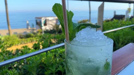 Delicious-fresh-cold-mojito-cocktail-at-a-beautiful-beach-bar-with-sea-view,-refreshing-drink,-enjoying-summer-with-dreamy-holiday-in-Marbella-Spain,-vacation-destination,-4K-shot