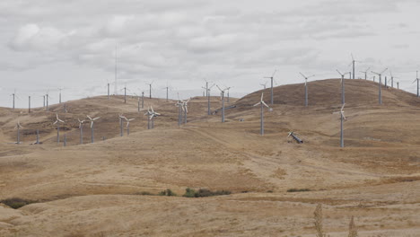 A-large-group-of-windmills-not-turning,-some-broken,-on-dry-hill-on-cloudy-day
