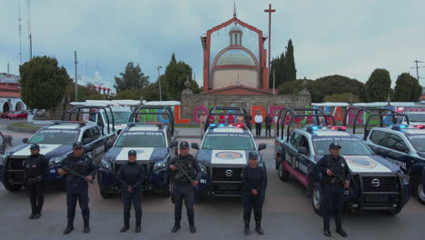 Vertical-pan-of-7-police-pick-ups-and-2-ambulances-with-armed-officers-of-the-Police-Department-lined-up-and-posing-in-front-of-their-cars-with-a-church-in-the-background