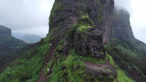 Drone-shot-revealing-landscape-of-ancient-Harihar-Fort-with-tourists-climbing-down-steep-and-vertical-stairs-during-monsoon-trek,-Harshewadi,-Trimbak