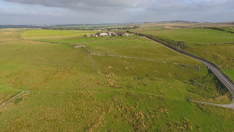 Drohnenaufnahmen-Vom-Podest,-Aufgenommen-Am-Frühen-Abend,-Zeigen-Die-Landschaft-Von-West-Yorkshire,-Darunter-Eine-Landstraße,-Einen-Bauernhof,-Felder-Und-Trockenmauern-Mit-Windkraftanlagen-In-Der-Ferne