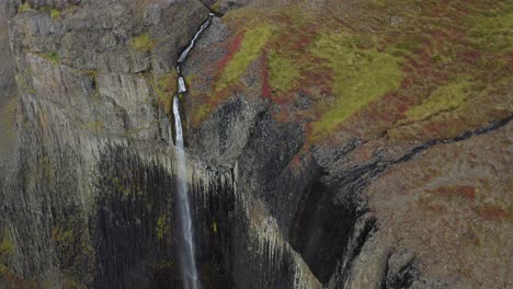 Die-Nach-Unten-Geneigte-Luftaufnahme-Zeigt-Einen-Atemberaubenden-Wasserfall,-Der-An-Einem-Bewölkten-Tag-Die-Isländische-Schlucht-Hinunterstürzt-–-Moosige-Vulkanlandschaft