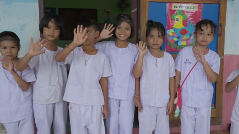 Thai-students,-positioned-in-front-of-their-classroom,-enthusiastically-wave-to-the-camera,-radiating-warmth-and-the-joy-of-learning