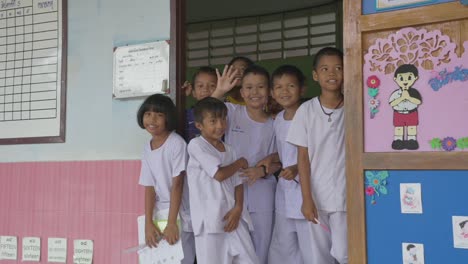 En-La-Puerta,-Los-Niños-Tailandeses-Se-Despiden-Con-La-Mano-Y-Sus-Sonrisas-Irradian-Calidez-Mientras-Se-Despiden,-Creando-Un-Momento-Conmovedor-De-Conexión-Compartida.