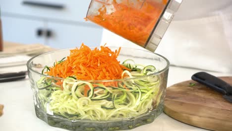 Älterer-Mann-Fügt-Zerkleinerte-Karotten-Zu-Spiralförmigem-Salat-Hinzu,-Um-Zucchini-Nudeln-Zuzubereiten,-Die-Zu-Einer-Schüssel-Mit-Gesunder-Veganer-Vegetarischer-Lebensweise-Und-Entgiftung-Beitragen