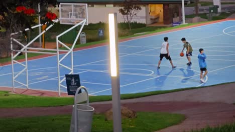 Teen-tries-but-fails-and-falls-while-playing-a-dusk-basketball-game-at-a-public-court-with-two-players,-a-trash-bin,-and-a-lamp-post-in-a-well-maintained-park