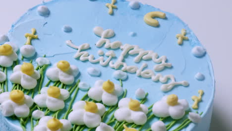 A-close-up-shot-of-a-birthday-cake-with-blue-creamy-frosting-and-decorated-with-white-and-yellow-flowers,-with-green-trimmings