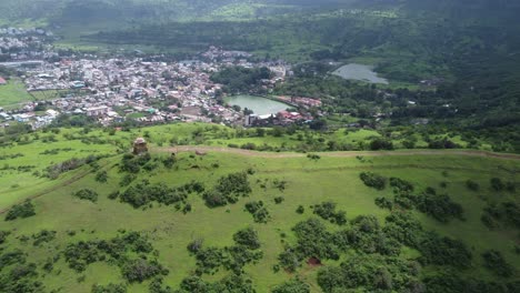 Aerial-view-of-the-Lord-Shiva-temple-at-the-edge-of-the-mountain-top-with-the-view-of-Trimbkeshwar-town-and-Ahilya-Dam-in-the-background-surrounded-by-lush-greenery,-Nashik,-Maharashtra,-India