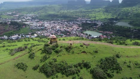 Drone-circling-around-Hindu-temple-at-the-edge-of-the-mountain-top-with-the-view-of-Trimbkeshwar-town,-Ahilya-Dam-and-Trimbak-falls-in-the-background-surrounded-by-lush-greenery,-Nashik,-India