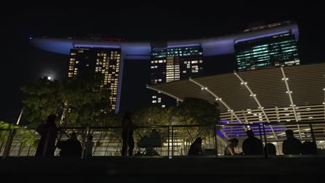 Blick-Von-Der-Promenade-Aus-Auf-Das-Beleuchtete-Marina-Bay-Sands-Hotel-In-Singapur-Bei-Nacht-Mit-Der-Silhouette-Von-Menschen-Neben-Geländern